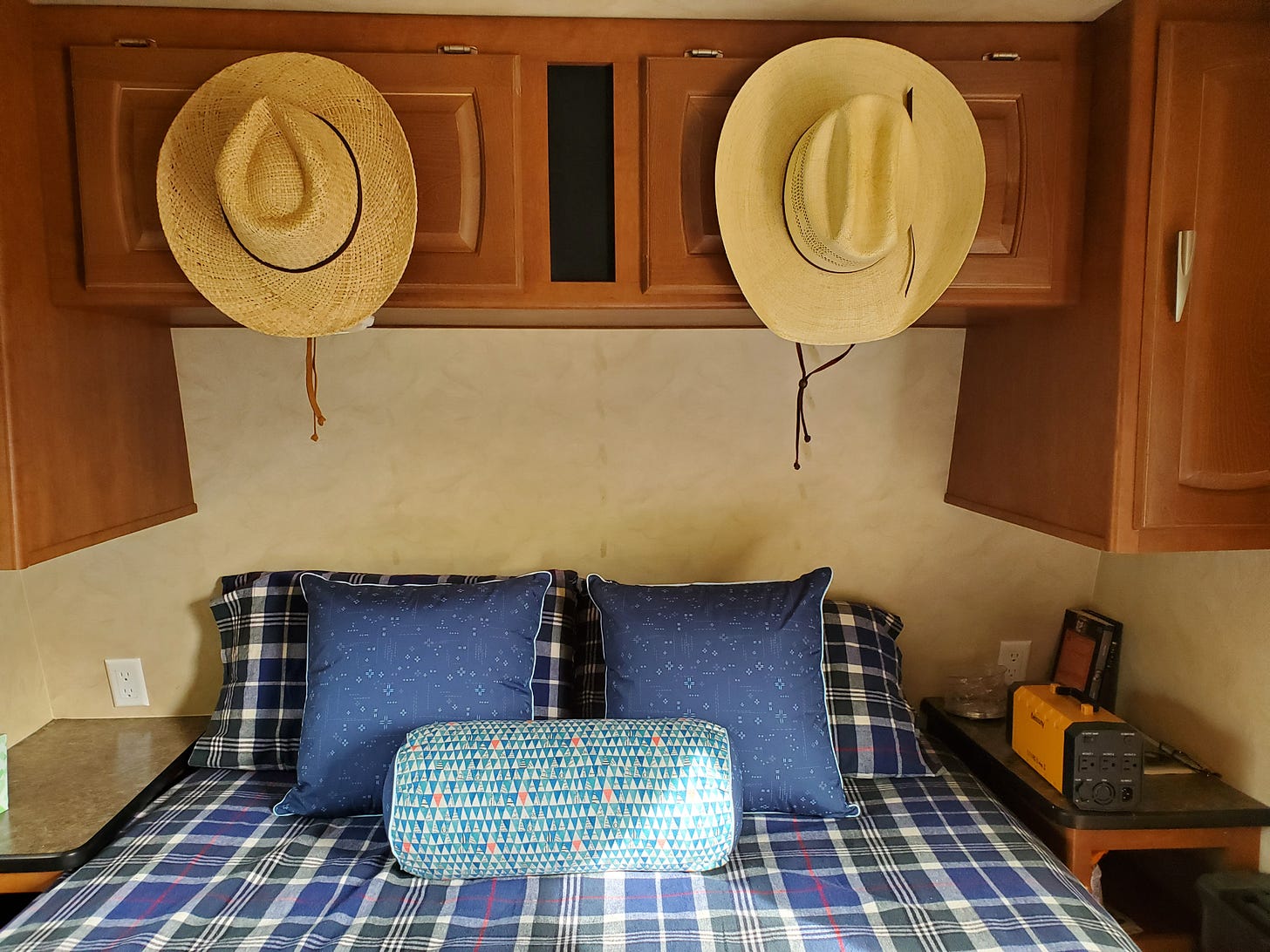 Interior shot of the travel trailer showing a queen bed with plaid sheets and pillows in various blue fabrics. Above the bed two cowboy hats hang his and hers style above from the upper cabinet.