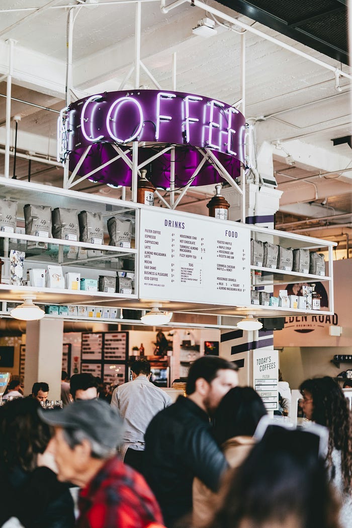 A big neon ‘coffee’ sign over the menu at a small coffee shop full of customers. Retail workers face customer abuse every day, and it’s very stressful.