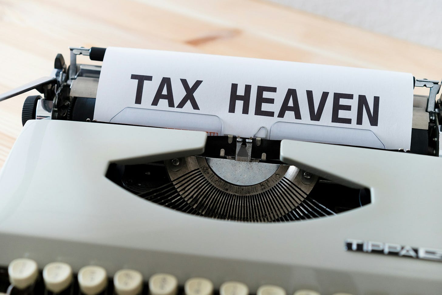 Close-up image of a white typewriter with a sheet of paper inserted that has "TAX HEAVEN" typed in large, bold black letters. The typewriter appears to be vintage style with its metal typebars visible beneath the paper and traditional keyboard keys visible at the bottom of the frame. The typewriter sits on a light wooden surface.