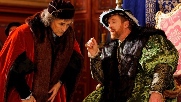 A man with a red beard dressed in opulent, green Tudor costume trimmed with fur sits on a throne and speaks to a hunched-over man in a red robe also trimmed with fur