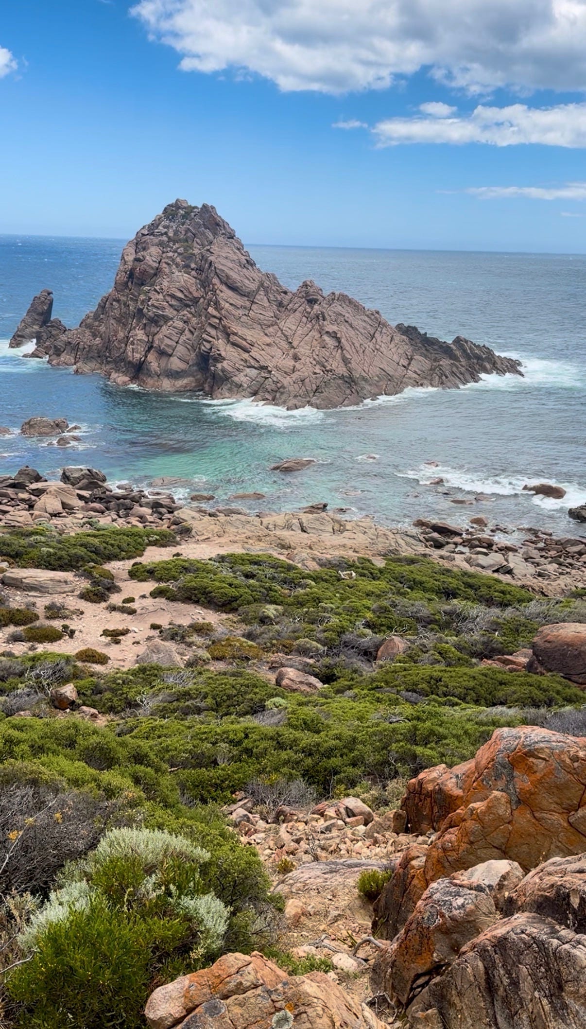 Closer view of Sugarloaf Rock from the shoreline, with rocky foreground, low coastal scrub, turquoise water, and the ocean stretching beyond the rock formation.