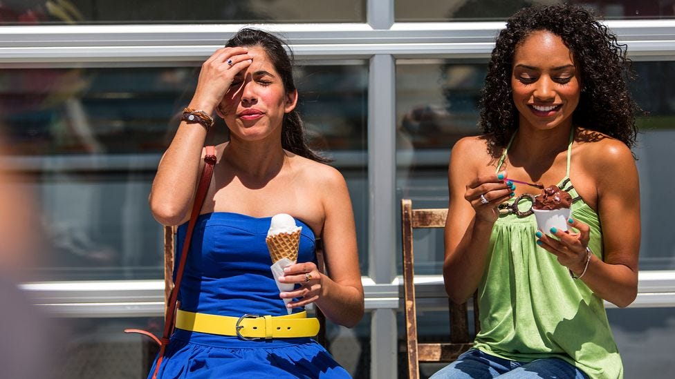 Two adult women sit outside in the sun against a windowed wall while eating ice cream. One woman holds her right hand up to her forehead while pursing her lips. (Credit: Getty Images)