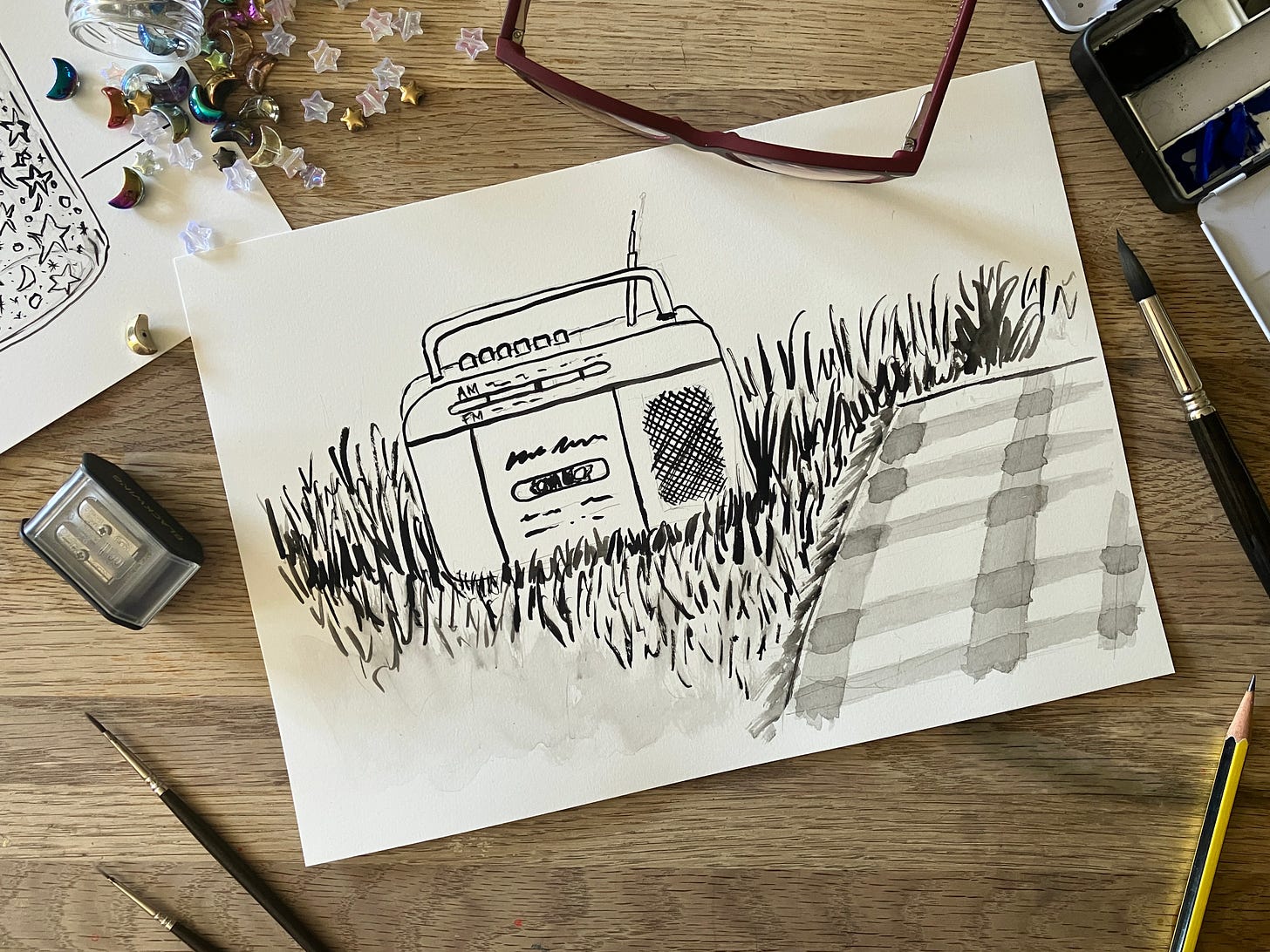 Painting of a cassette player on grassy ground next to a picnic rug sits atop a table surrounded by art materials