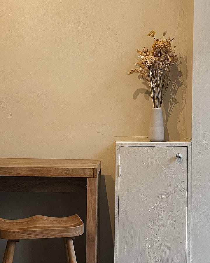 Image 1: Cosy window seating with cushions, a small wooden table holding a cup of coffee and a glass of water. / Image 2: Scandi-inspired seating with high stools, wooden furniture, and a vase of dried flowers in soft beige and yellow tones.