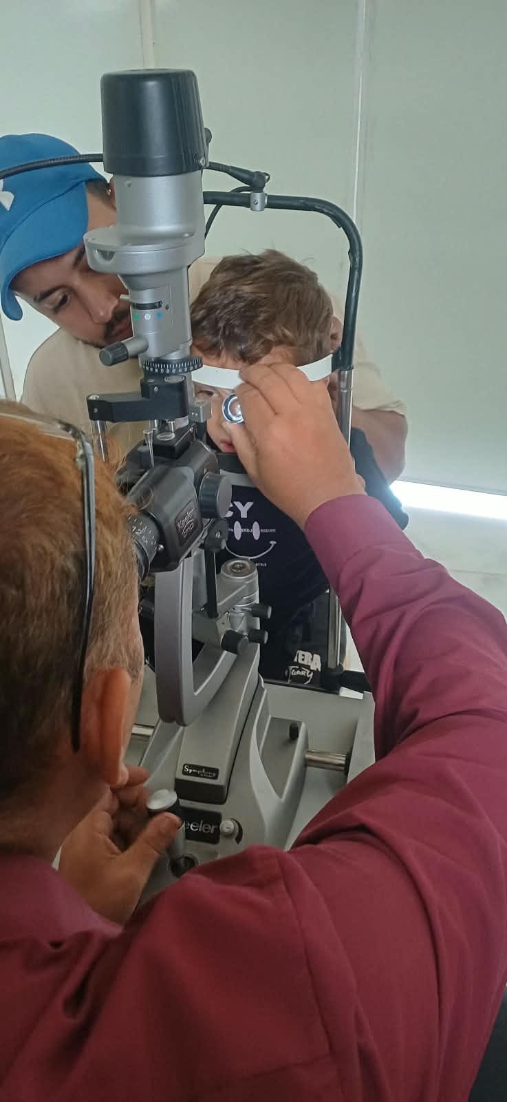 A young child undergoes an eye examination using a slit-lamp microscope while an eye care professional adjusts the instrument. A second adult assists the child during the examination in a clinical setting.