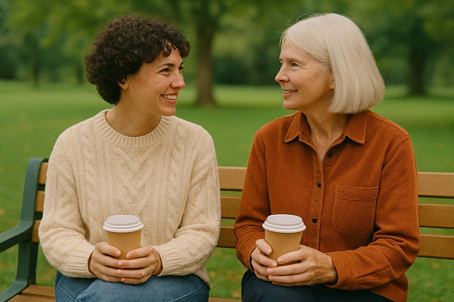Two people sit on a wooden park bench holding coffee cups, turned toward each other and smiling as they talk. Trees and green grass fill the background, creating a calm, everyday moment of connection and companionship.