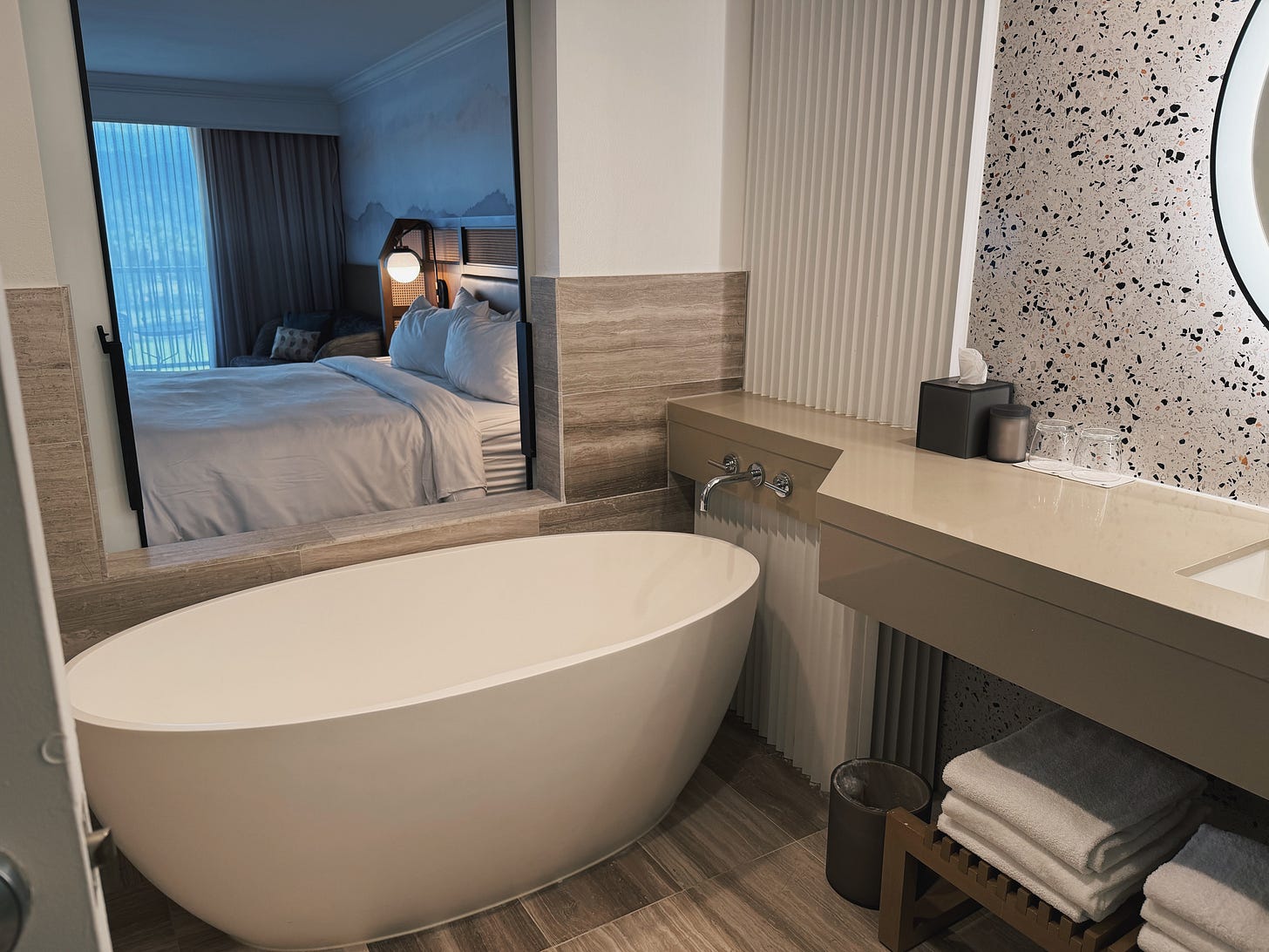 A freestanding soaking tub in the renovated bathroom at JW Marriott Desert Springs, with the bedroom visible through an open interior window. A freestanding soaking tub in the renovated bathroom at JW Marriott Desert Springs, with the bedroom visible through an open interior window.