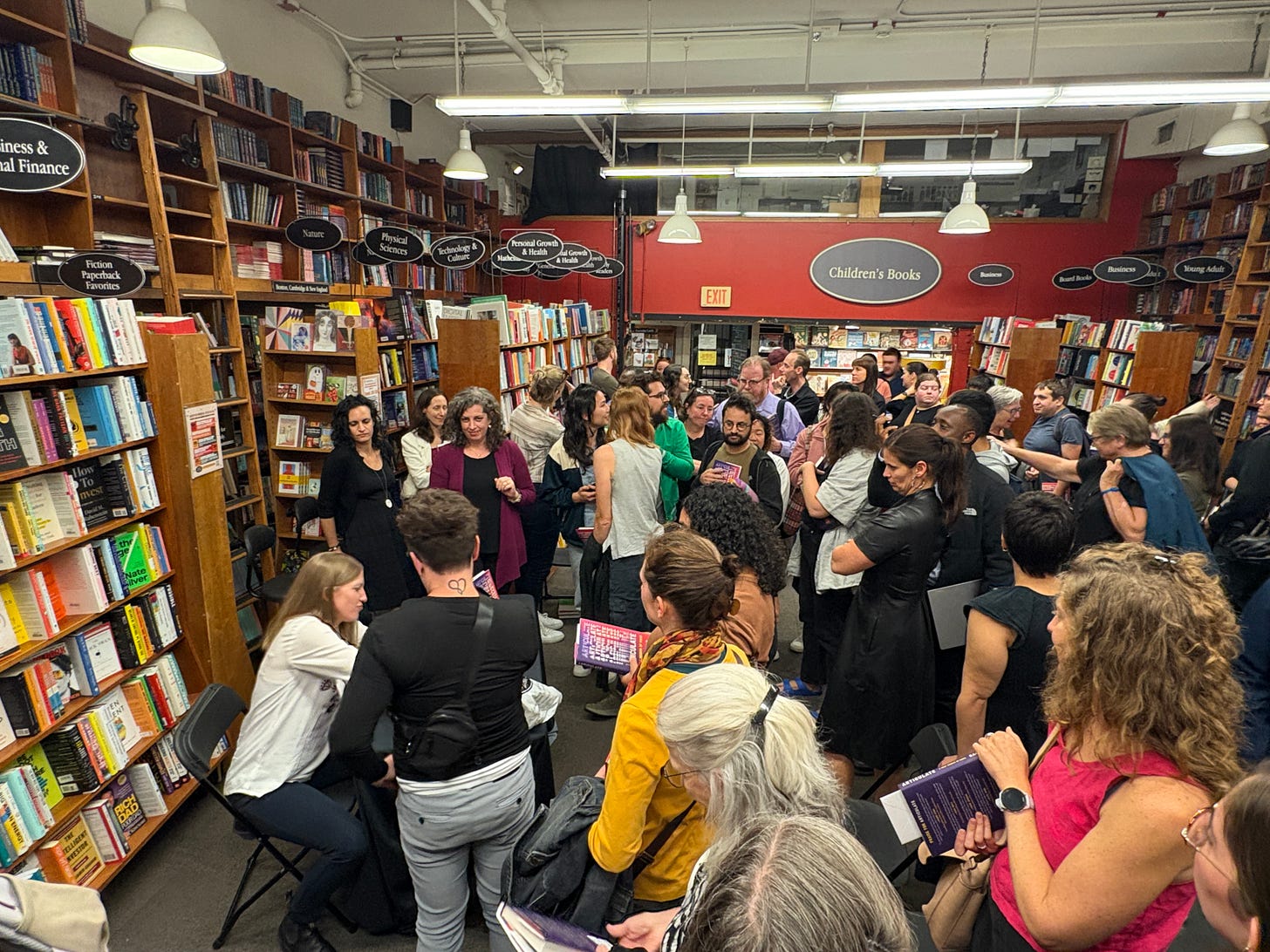 Photo of the crowd milling around the room and lined up for a book signing after my book event in Harvard Bookstore. Many people are clustered in a room lined with bookshelves, and I'm sitting in one corner signing someone's book at a table.