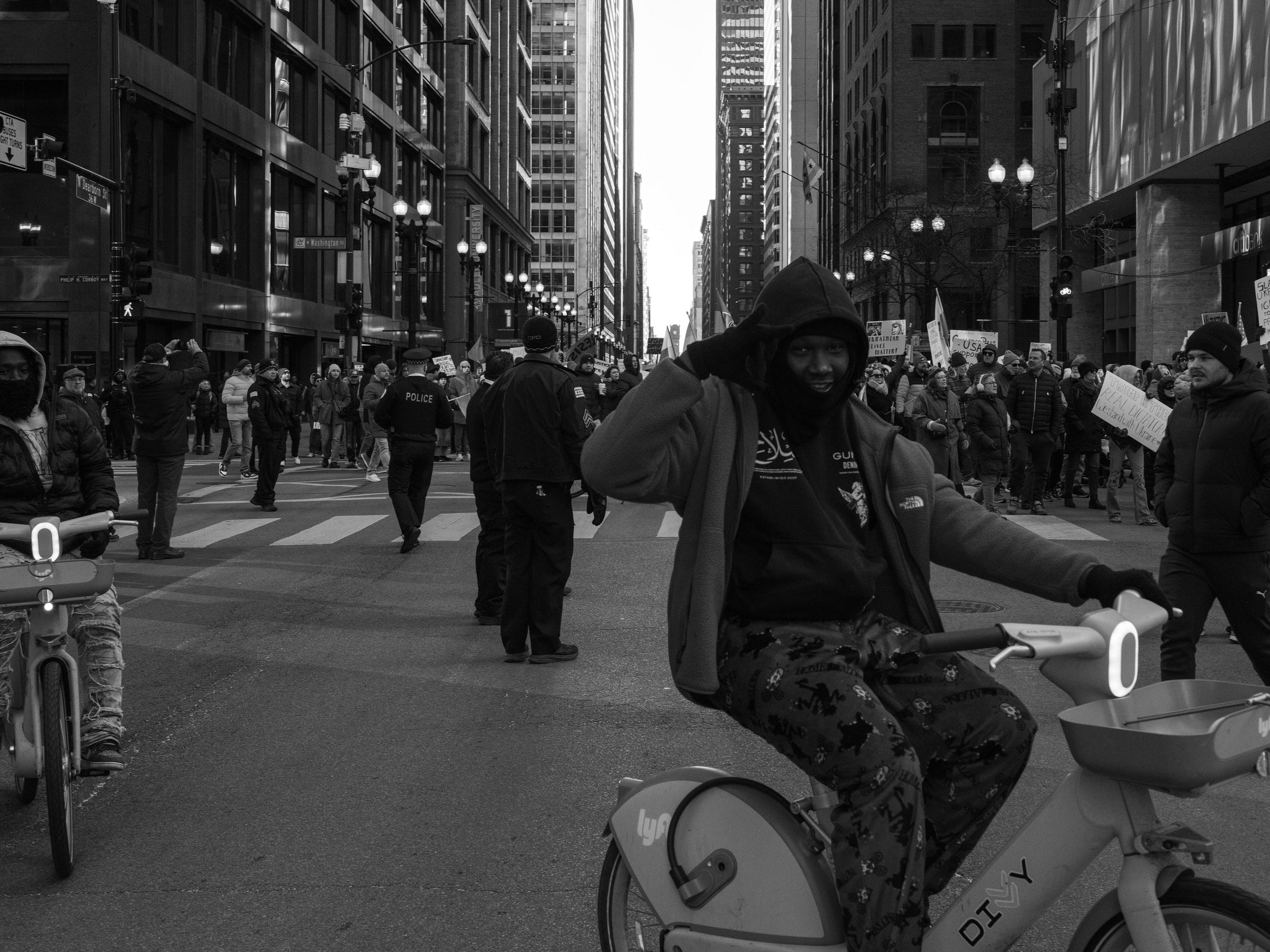 A black and white photograph captures a moment at a protest in a city intersection. In the foreground, a young man in a hoodie and patterned pants rides a Divvy bike, saluting the camera with a playful expression. Another cyclist wearing a mask rides beside him. In the background, police officers monitor a large crowd of protesters holding signs, while tall buildings and streetlights frame the urban scene. The contrast between the demonstrators and the cyclists adds a dynamic layer to the image. A black and white photograph captures a moment at a protest in a city intersection. In the foreground, a young man in a hoodie and patterned pants rides a Divvy bike, saluting the camera with a playful expression. Another cyclist wearing a mask rides beside him. In the background, police officers monitor a large crowd of protesters holding signs, while tall buildings and streetlights frame the urban scene. The contrast between the demonstrators and the cyclists adds a dynamic layer to the image.