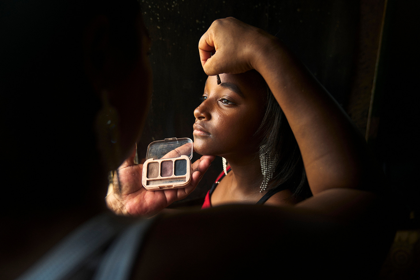A woman applies makeup to another womans face, holding an eyeshadow palette in one hand. The woman receiving makeup has long dark hair and wears sparkling earrings, gazing up while being carefully attended to.