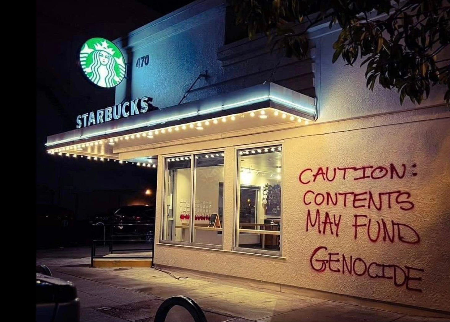 Exterior of a Starbucks store at night with the text 'CAUTION: CONTENTS MAY FUND GENOCIDE' spray-painted in red on the wall next to the entrance. Exterior of a Starbucks store at night with the text 'CAUTION: CONTENTS MAY FUND GENOCIDE' spray-painted in red on the wall next to the entrance.