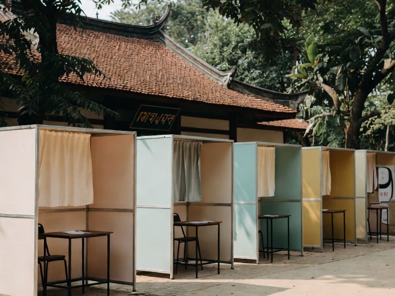 A photograph of multiple polling booths in an outdoor Asian location, arranged in a row with a traditional building in the background. A photograph of multiple polling booths in an outdoor Asian location, arranged in a row with a traditional building in the background.