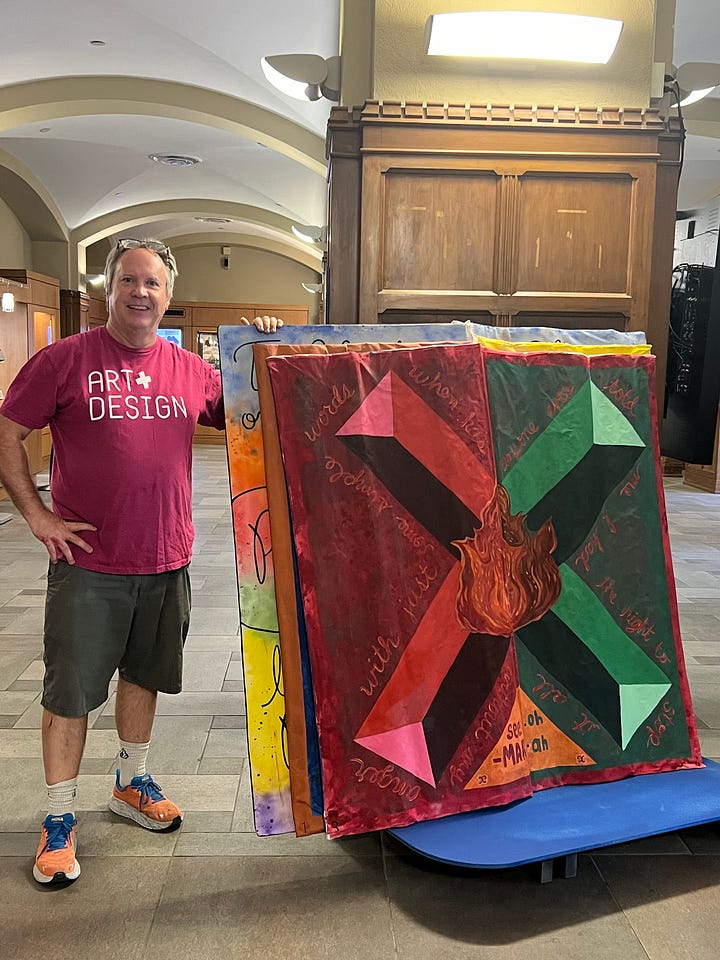 A big book with colorful pages sits on a display stand in front of a pair of windows. A painted, oversized book sits on a green display stand with library shelves visible behind it. A man stands next to an oversized book that is painted in red and green.