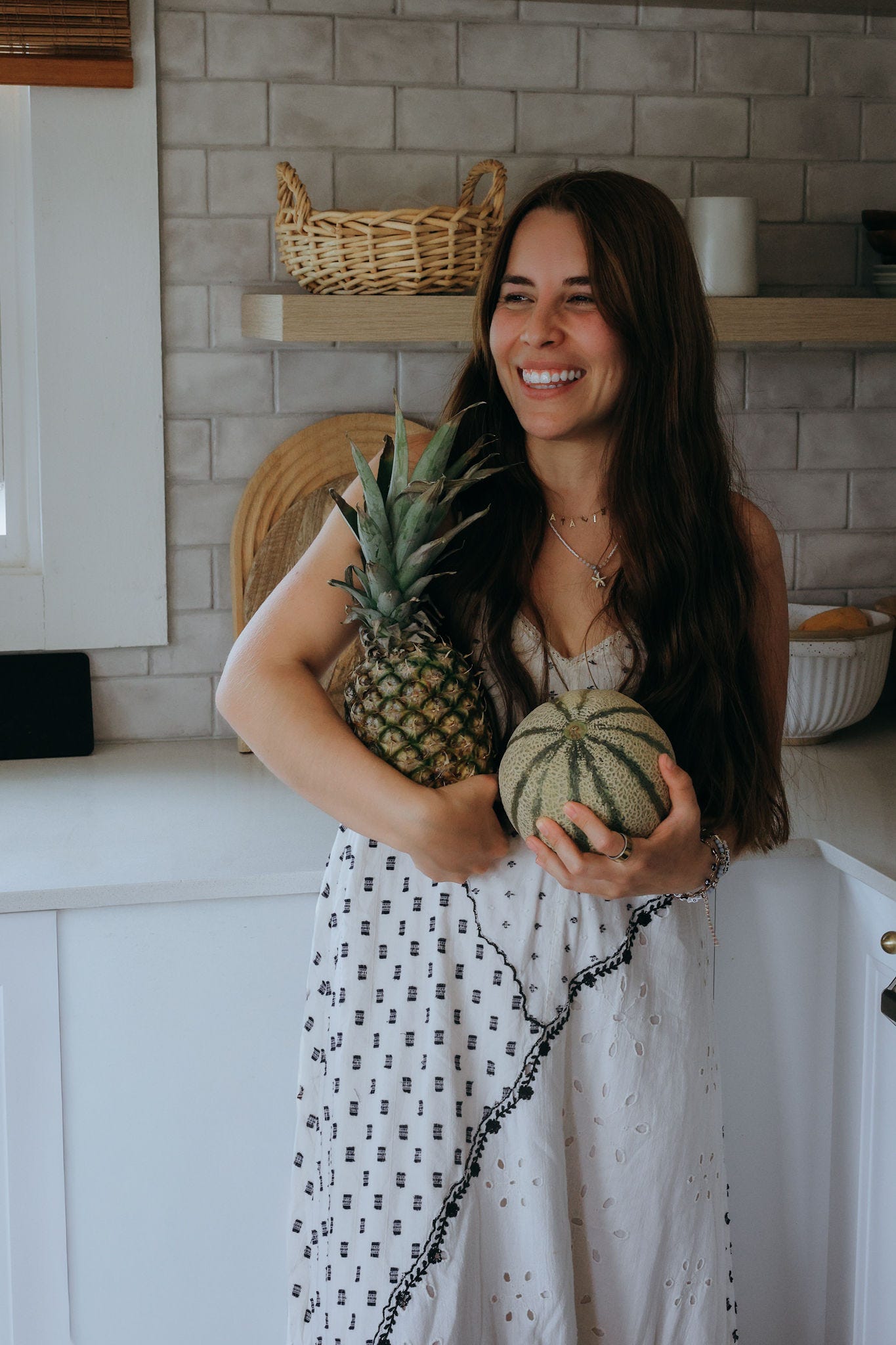 a woman with long dark hair and print white dress standing in her kitchen holding a pineapple and melon a woman with long dark hair and print white dress standing in her kitchen holding a pineapple and melon