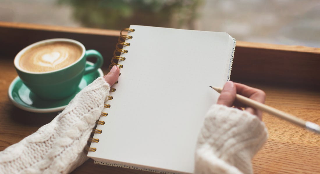 Two hands with cream knit sleeves holding a plain notebook in front of a wooden desk with a coffee cup on it Two hands with cream knit sleeves holding a plain notebook in front of a wooden desk with a coffee cup on it
