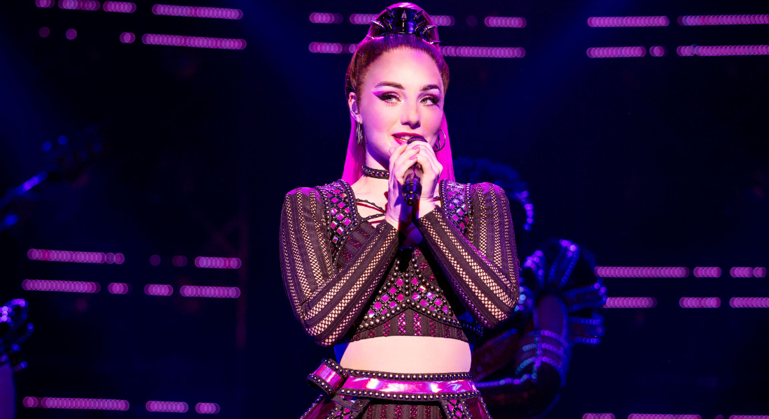 A woman wearing a black mesh crop too and skirt singing into a microphone, with purple lights around her