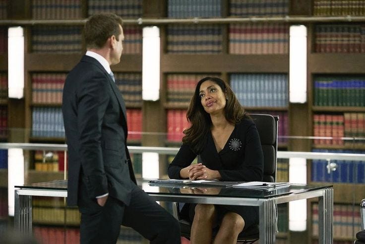 This may contain: a woman sitting at a desk talking to a man in front of bookshelves