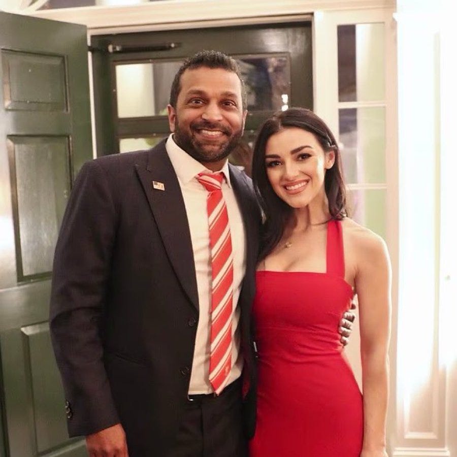 Elijah Schaffer stands next to a woman in a red dress. He wears a dark suit, white shirt, and red striped tie. They are positioned in front of a green door with white trim, smiling at the camera.
