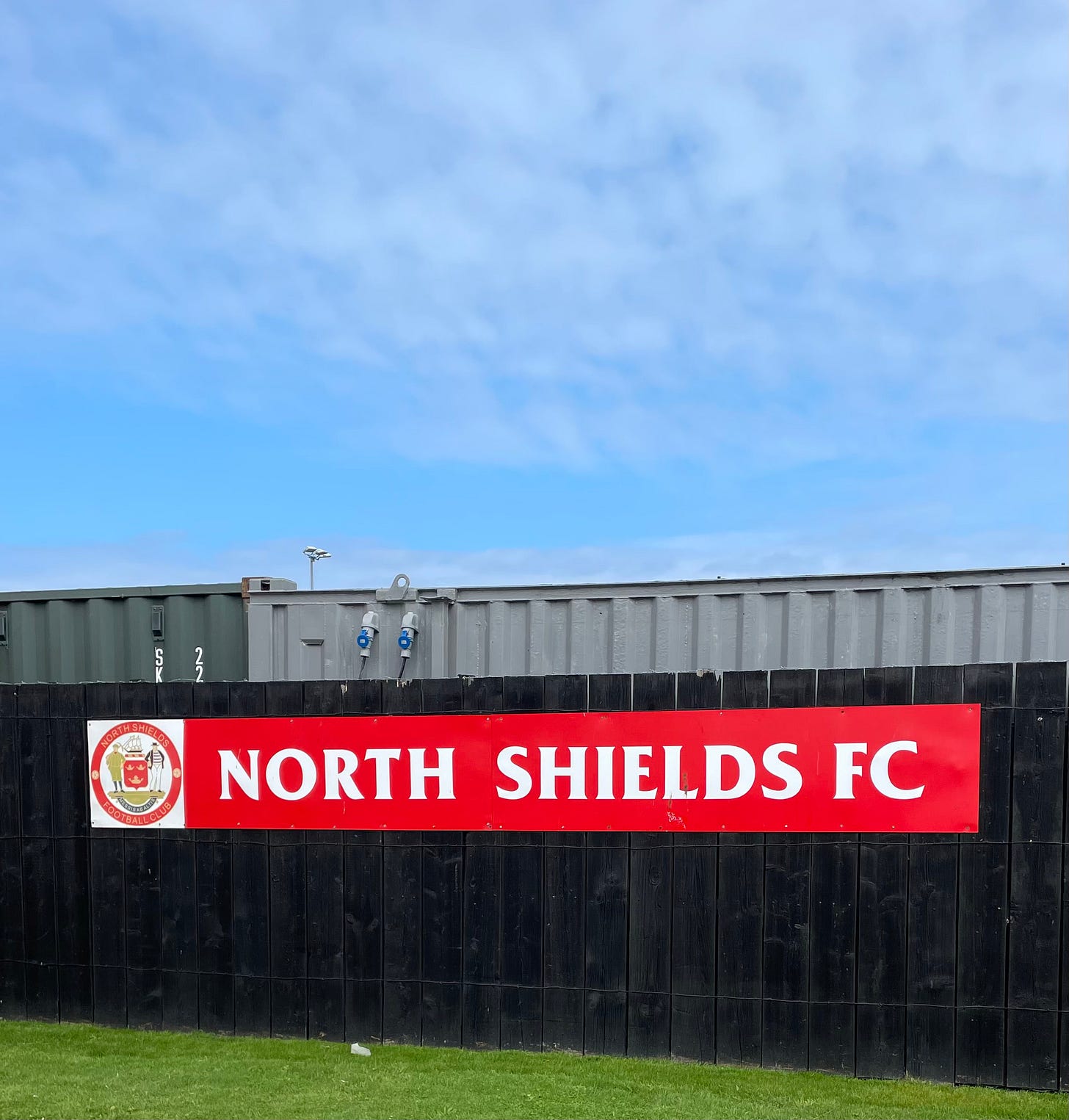 A blue sky, a grey fence and a red signboard welcome us to North Shields FC