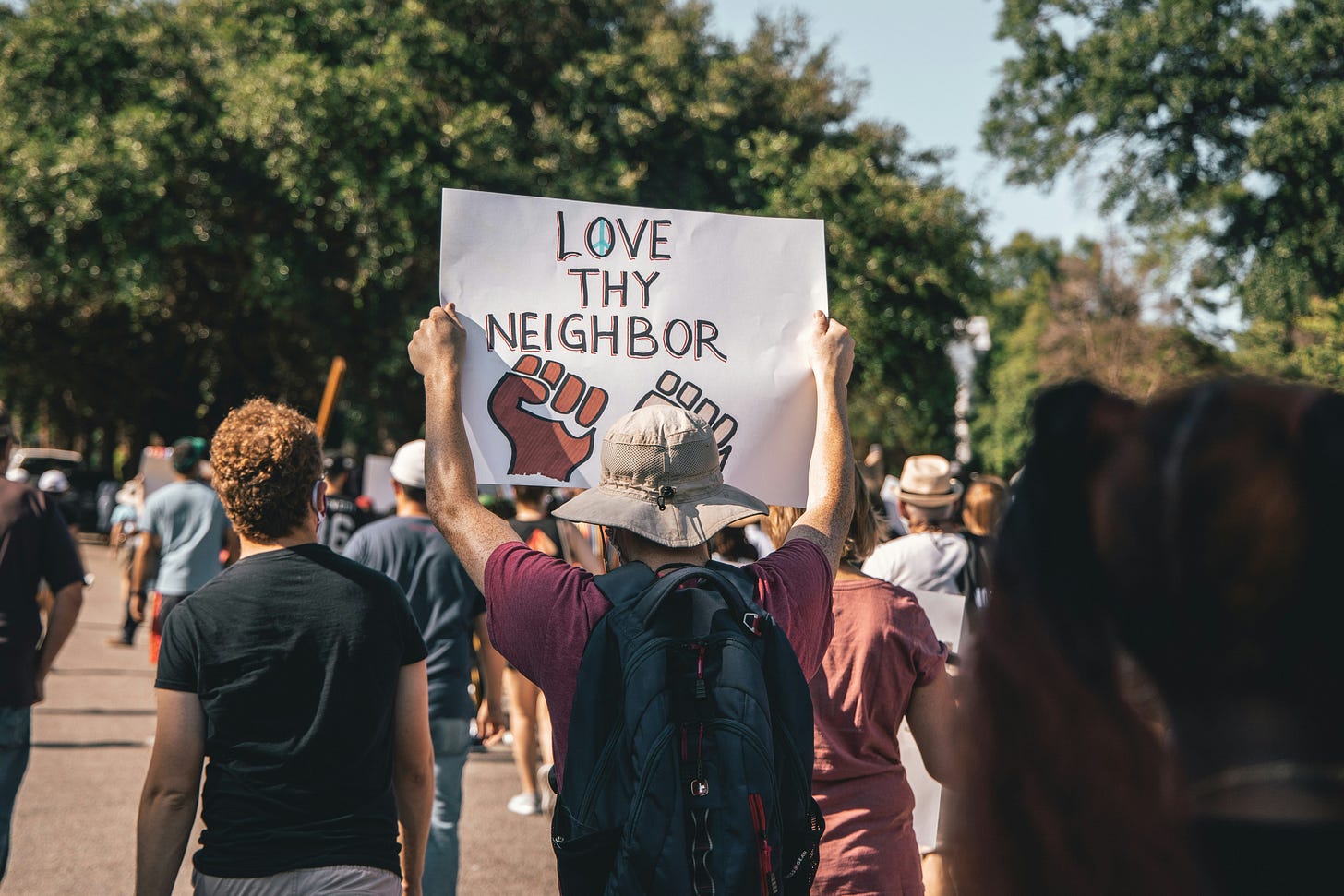 A person in a hat holds a "Love Thy Neighbor" sign with a raised fist. They walk with a diverse crowd in a peaceful protest. Sunny day, green trees.