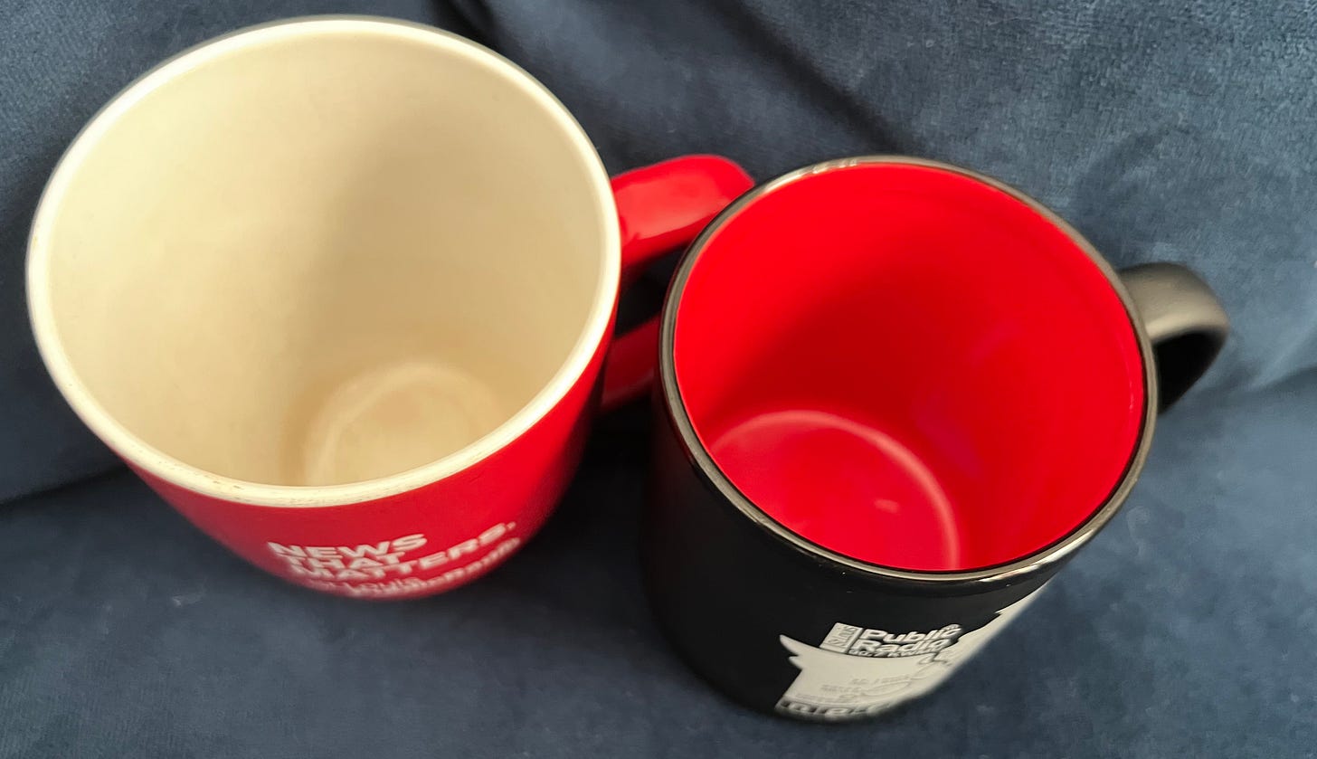 Image is of a view from above of two coffee ups. A red one has a white interior that is stained with coffee. A black one has a red interior with no visible stains. Both are coffee mugs with a logo of a local National Public Radio station.