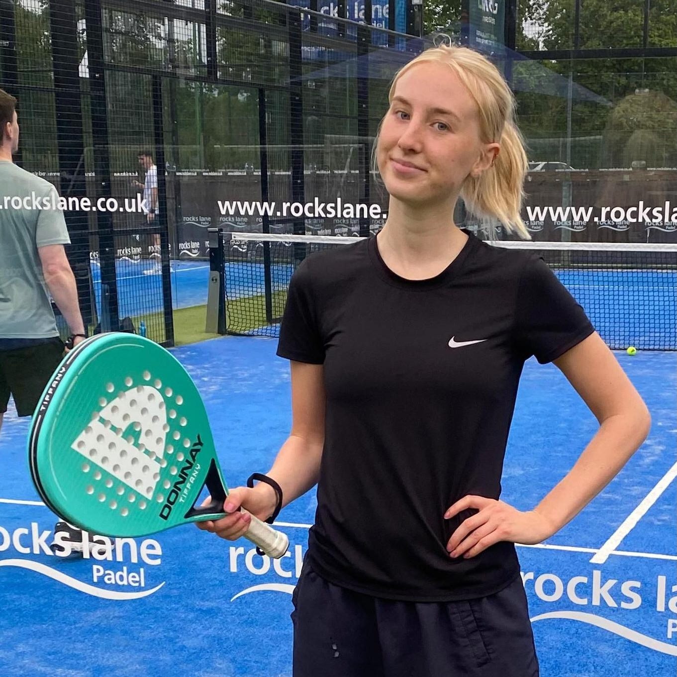 Rosie is standing in front of a blue astro padel court. She is wearing a black t-shirt and matching tracksuit bottoms and we can only see her from the waist up. She is holding a turquoise padel racquet in one hand and the other hand is on her hip. Rosie has shot blonde hair, which has been tied back into a low ponytail. She is smiling into the camera with no teeth. Behind her are two men playing on the court, who can be seen through the glass walls. There is white branding written on the glass. Rosie is standing in front of a blue astro padel court. She is wearing a black t-shirt and matching tracksuit bottoms and we can only see her from the waist up. She is holding a turquoise padel racquet in one hand and the other hand is on her hip. Rosie has shot blonde hair, which has been tied back into a low ponytail. She is smiling into the camera with no teeth. Behind her are two men playing on the court, who can be seen through the glass walls. There is white branding written on the glass.