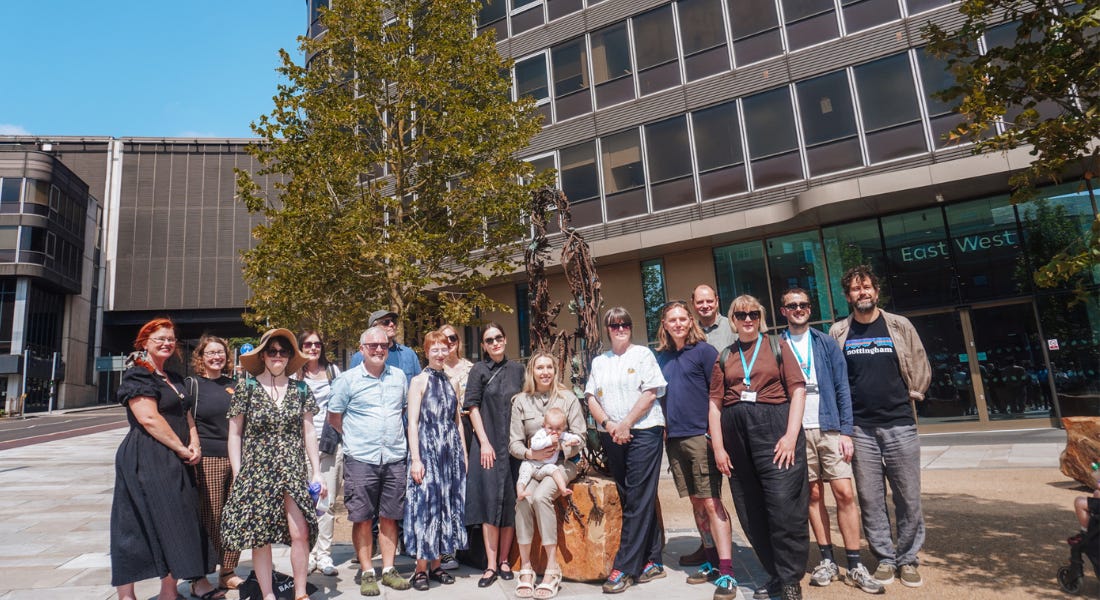 A group of people crowded around the Maid Marian statue with a tree and buildings behind them A group of people crowded around the Maid Marian statue with a tree and buildings behind them