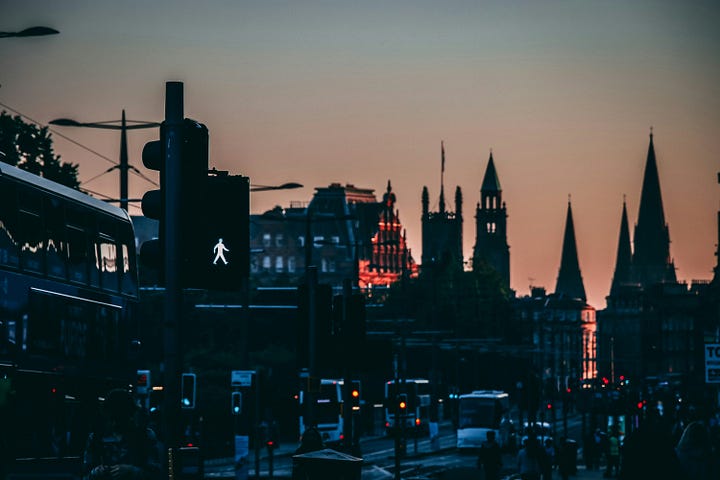 In the first picture a sunset landscape, th Edimburgh Castle siluetted in the background against the sky, red sunset light reflecting on its walls, in the foreground, in shadow, a busy street. A pedestrian crossing, cars and busses. In the second picture In the foreground, an ornate letter with elaborate calligraphy lies on a polished dark wood desk. Beyond the window, the amber lights of Edinburgh's Old Town blur through heavy rain.