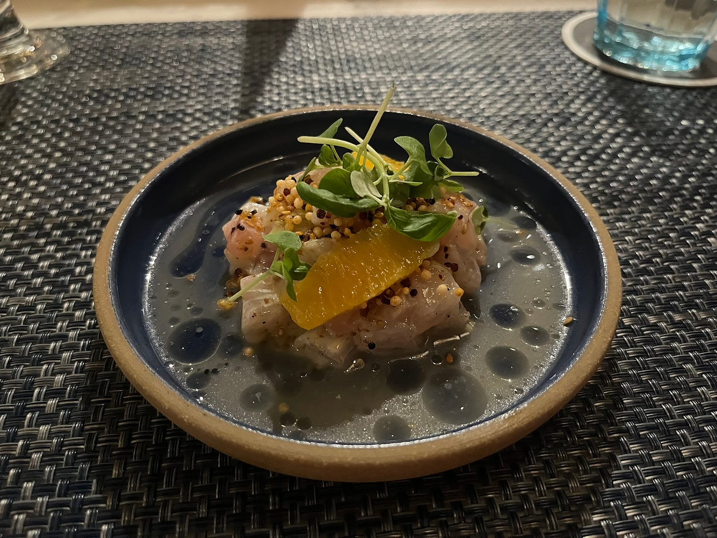 Pinkish white pieces of fish, served with small orange slices, white and brown seeds, and light greens, on a small round plate