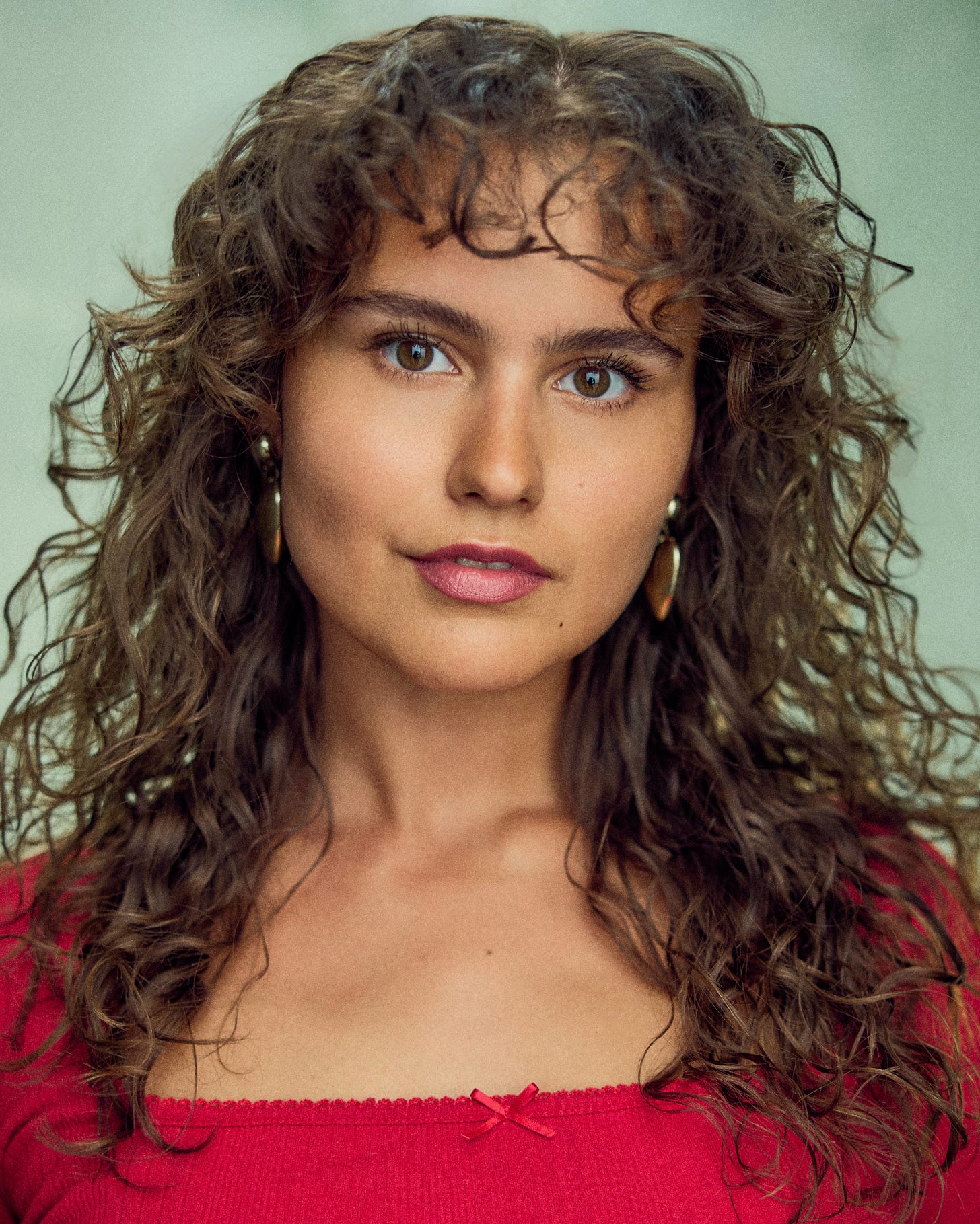 Head-and-shoulders portrait of Boo Miller looking directly at the camera, with long curly brown hair, gold hoop earrings and a red top against a pale green background. Head-and-shoulders portrait of Boo Miller looking directly at the camera, with long curly brown hair, gold hoop earrings and a red top against a pale green background.