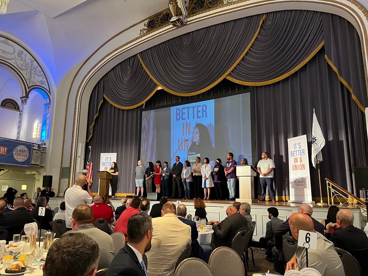 About a dozen people stand on the ballroom stage, with a person in a SAG-AFTRA shirt speaking at the microphone in front of a banner that says "It's Better In A Union"
