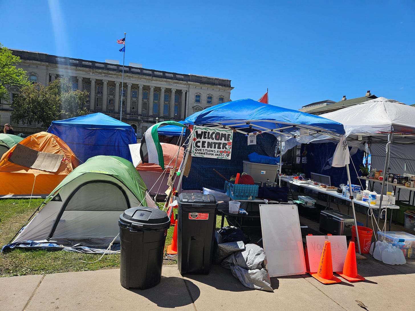 Camping tents are clustered along a grassy area, with two awning-style tents at the center, one with a "Welcome Center" sign hanging in it, and the other filled with food for distributing. A Palestinian flag waves from the side of one of the tents.