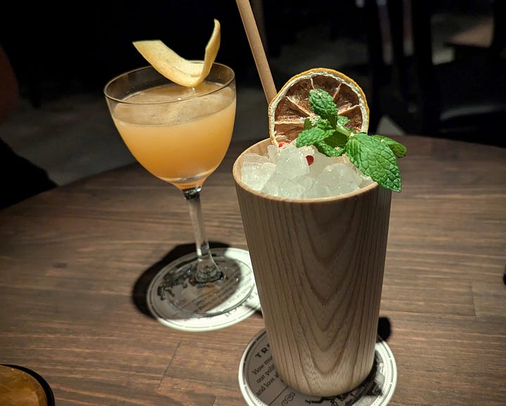 Two cocktails on a table. A nighttime bar scene with thick wooden ceiling beams and dim lighting.
