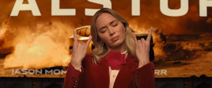 Emily Blunt dressed in a bold red suit flashing devil horns with both hands, in front of a stylized film poster for "Metalstorm."