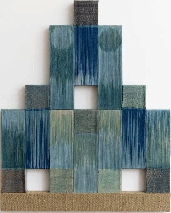 Carolina Jiménez, Spring Rain, 2026. A woven wall piece stepped like a pyramid, built from blocks of indigo, teal and blue-grey thread, with three small square openings and a band of pale natural wood at the base. Carolina Jiménez, Spring Rain, 2026. A woven wall piece stepped like a pyramid, built from blocks of indigo, teal and blue-grey thread, with three small square openings and a band of pale natural wood at the base.