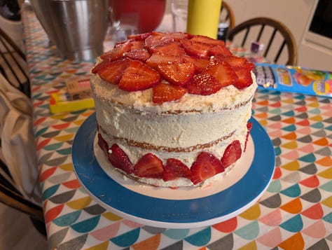 A Japanese Strawberry Shortcake triple layer cake with cream frosting and strawberry decoration, a club sandwich full of cooked breakfast items, and an adjustable angle art desk