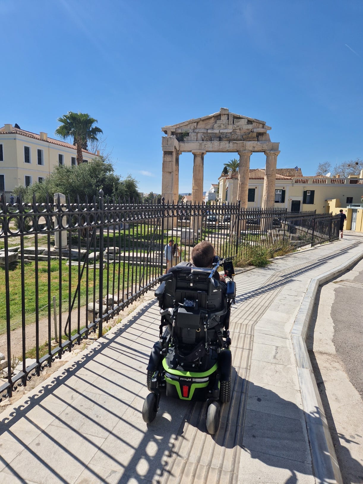 I am rolling on a sidewalk over a tactile path. In the background we see the Gate of Athena Archegetis and other buildings.