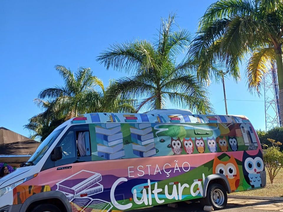 #descrevemospravocê  A imagem mostra uma van colorida com o letreiro “Estação Cultural” estampado em letras grandes na lateral. O veículo está estacionado sob um céu azul claro, com várias palmeiras ao fundo, sugerindo um dia ensolarado em uma cidade de clima tropical.  A lateral da van é ilustrada com elementos gráficos vibrantes. Na parte superior, há pilhas de livros e prateleiras com livros organizados. A parte central é decorada com desenhos de corujas de diferentes cores — rosa, laranja, branca, azul e marrom — representando sabedoria e diversidade. Também há árvores, folhas e uma estrada sinuosa que atravessa a paisagem ilustrada. Perto da traseira do veículo, aparece uma grande coruja com olhos azuis, que chama bastante atenção. À frente, sobre fundo roxo e rosa, estão dois livros abertos desenhados em branco.  O estilo visual da van é lúdico e acolhedor, com cores alegres e temática ligada à educação, leitura e cultura, sugerindo que o veículo funciona como biblioteca itinerante ou espaço cultural móvel.
