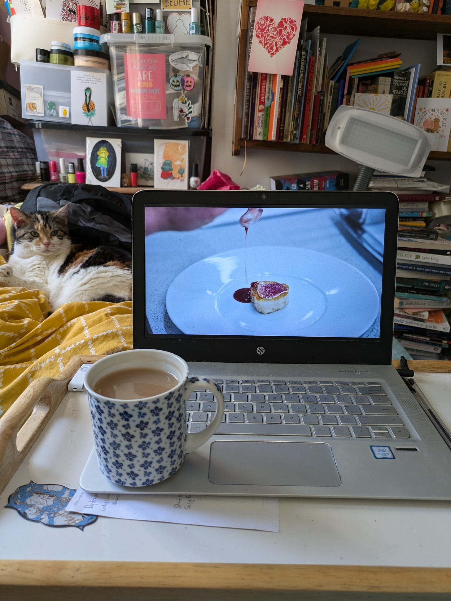 ID. A photo of a mug of tea balanced on a laptop on a tray table on a bed. On the screen is a still frame from s3 The Bear where a fine dining plate is being finished off with a drizzle. In the background you can see colourful bookshelves, storage boxes and a calico cat. ID. A photo of a mug of tea balanced on a laptop on a tray table on a bed. On the screen is a still frame from s3 The Bear where a fine dining plate is being finished off with a drizzle. In the background you can see colourful bookshelves, storage boxes and a calico cat.