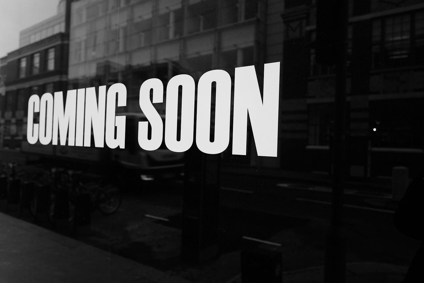 White large block capital letters spelling out COMING SOON along a window, with reflected buildings behind them. The photo is black and white. White large block capital letters spelling out COMING SOON along a window, with reflected buildings behind them. The photo is black and white.