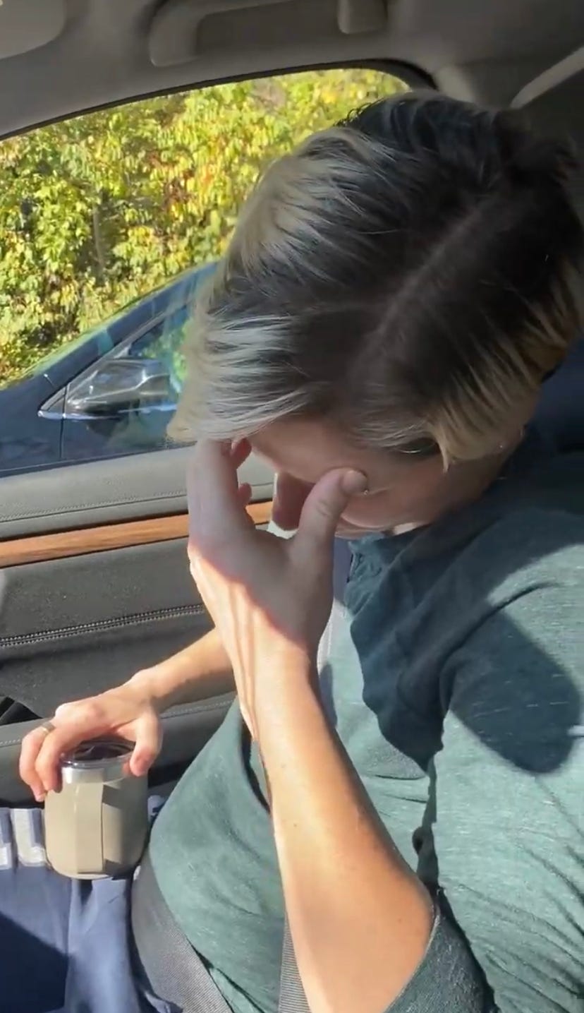 A person holding their head down with one hand, holding a cup of tea with another as they sit in the car. They have a long sleeved green shirt with blue sweatpants on. Their hair is short and has blonde highlights.