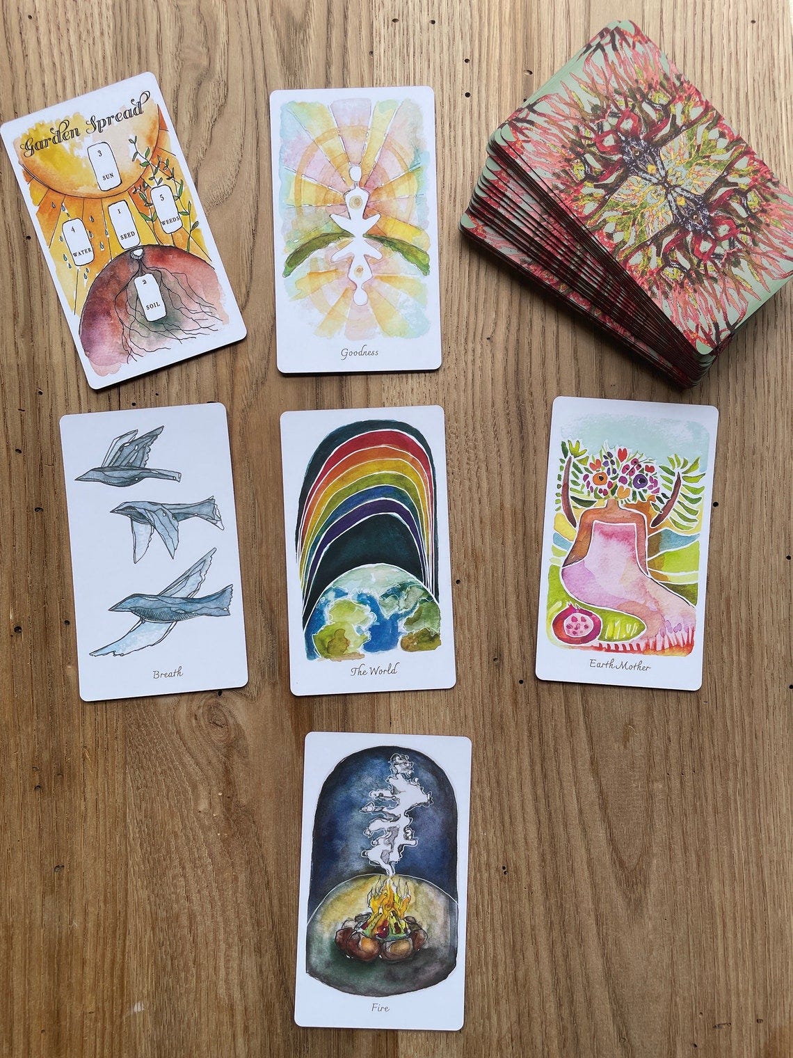 May include: A spread of tarot cards on a wooden surface. The cards are watercolor illustrations with a variety of colors and designs. The cards include 'Garden Spread', 'Goodness', 'Breath', 'The World', 'Earth Mother', and 'Fire'.