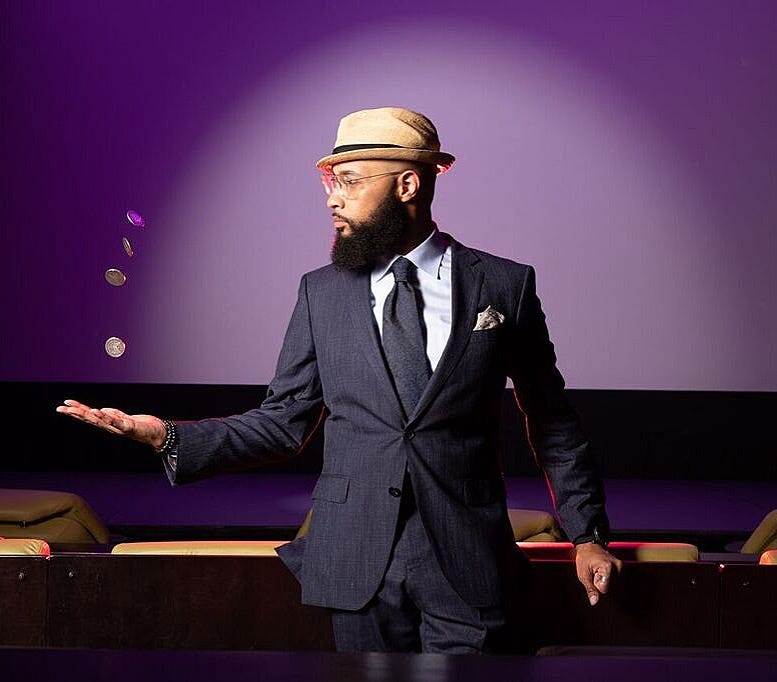 A bearded man in a suit and fedora looking at four coins suspended in the air above his outstretched hand against a purple backdrop. A bearded man in a suit and fedora looking at four coins suspended in the air above his outstretched hand against a purple backdrop.