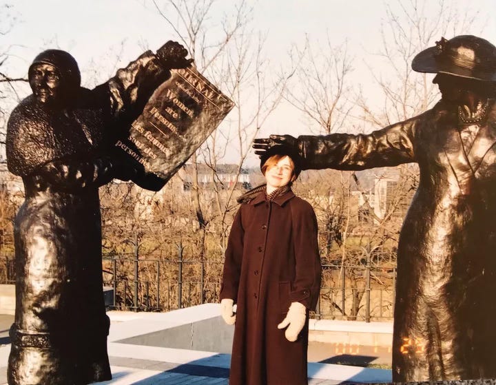 A young-adult me wearing the brown coat, standing between two statues of women in bronze, each wearing their own brown coat. One is holding a large bronze newspaper that reads “Women are Persons…Les femmes sont des personnes…” 