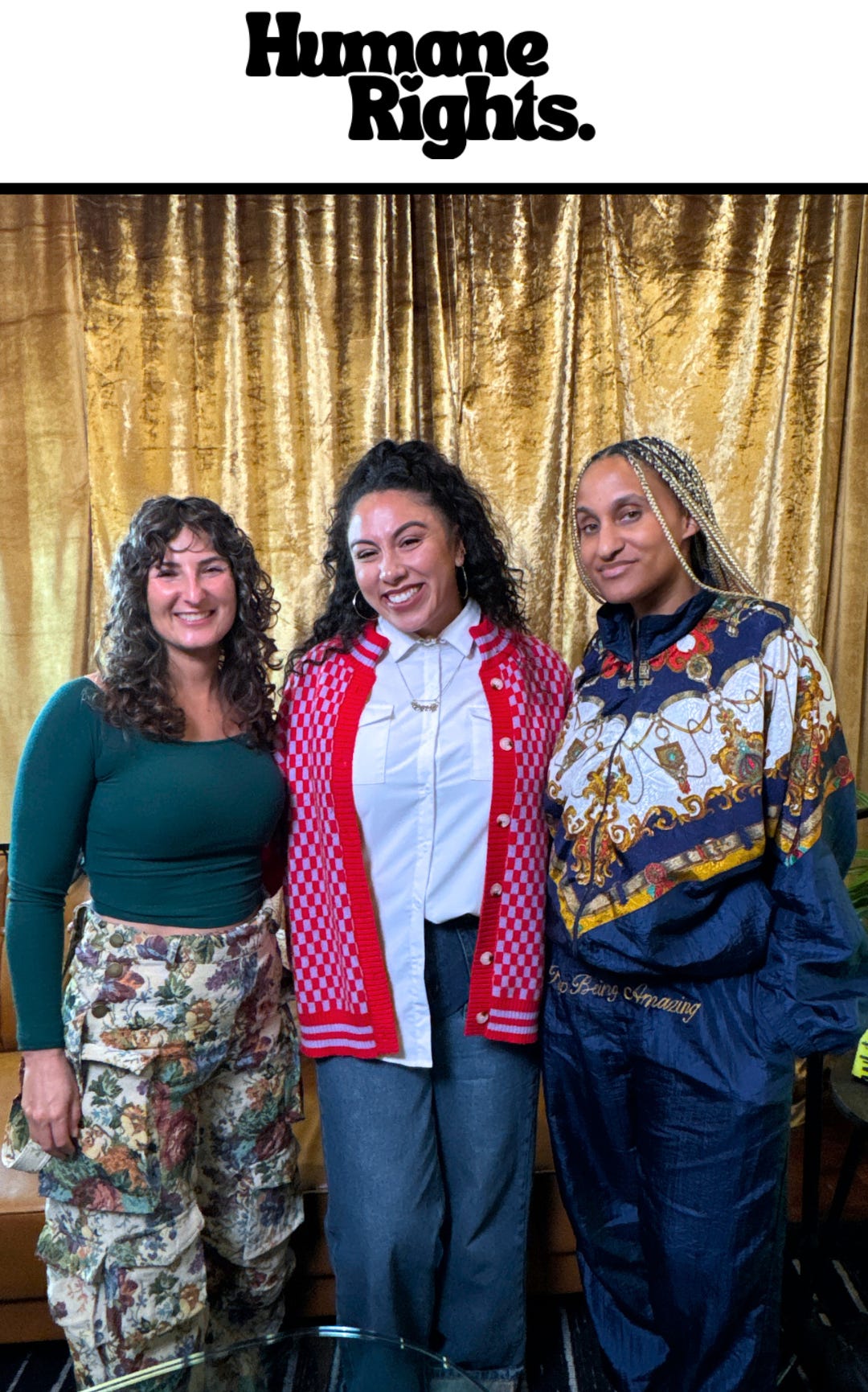 Dr. Cadet, who wears a vintage navy blue and ornate gold vintage tracksuit, stands next Grace, with long curls, a lavender and red cardigan and dark jeans, who stands next to Sam with dark curly hair, green top, and tan tapestry cargo pants in the Humane Rights Studio.