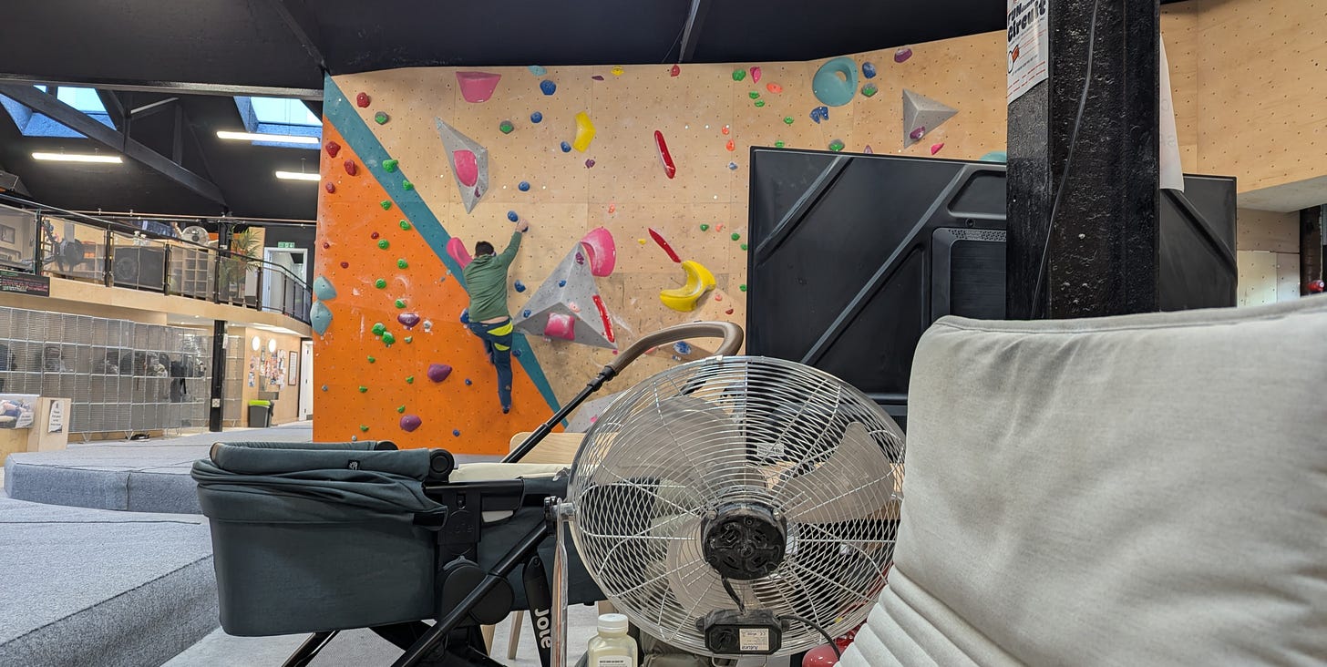 A pram in the foreground, inside of a climbing gym. In the background, a man is halfway up the climbing wall.