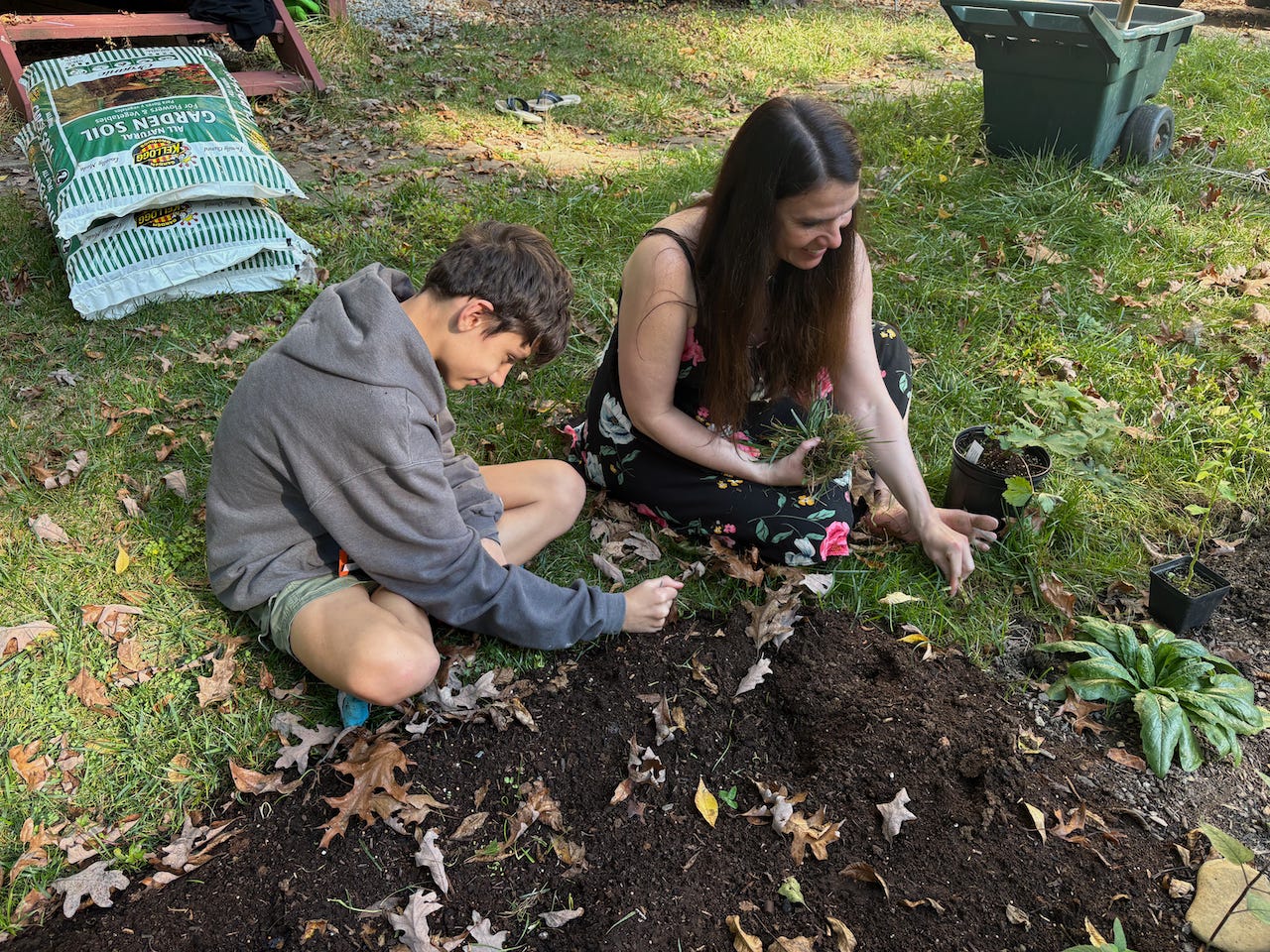 A boy and a woman sit cross-legged on the grass before a patch of earth with plants and fall leaves. They are pulling grass and smiling. Behind them there are earth bags and gardening tools. It is a sunny, early fall afternoon.