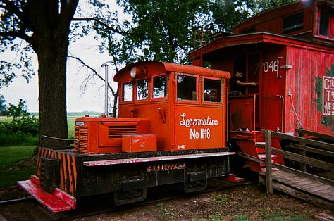 Red train engine with caboose. | An old typewriter used to relay messages to passing trains. | A white and yellow schoolhouse with an American flag.