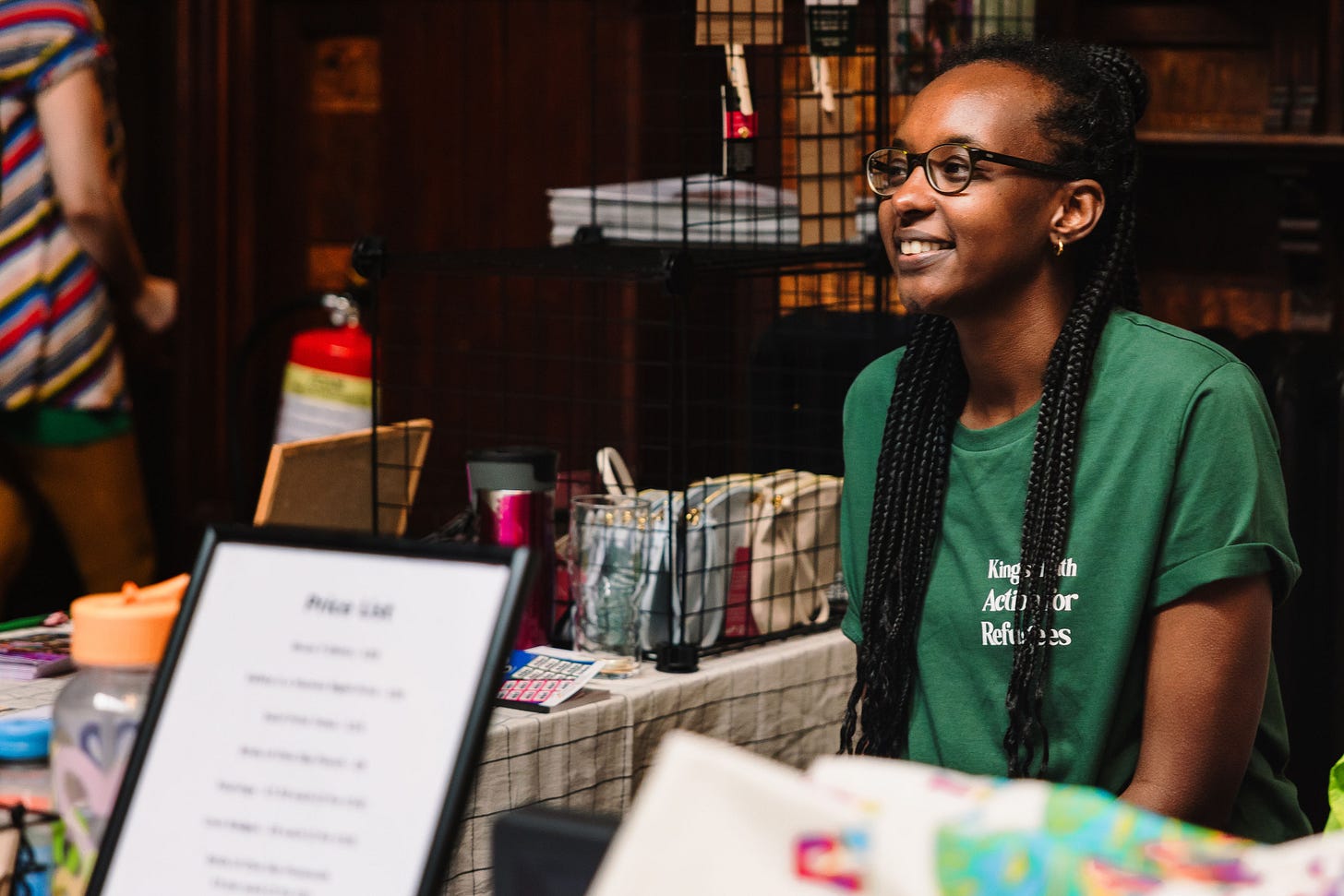 Female volunteer with long hair in a green t-shirt sits at the welcome desk and merchandise stall at KHARnival 2023