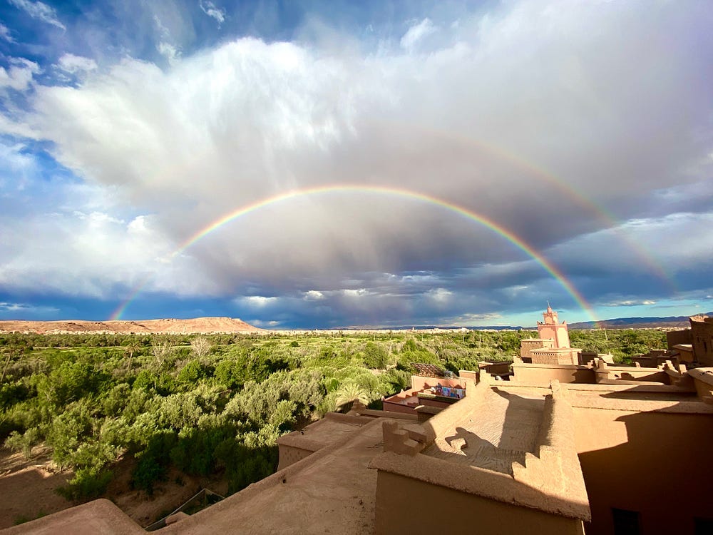A full rainbow arcs over a traditional Moroccan medina and extensive garden space beyond.